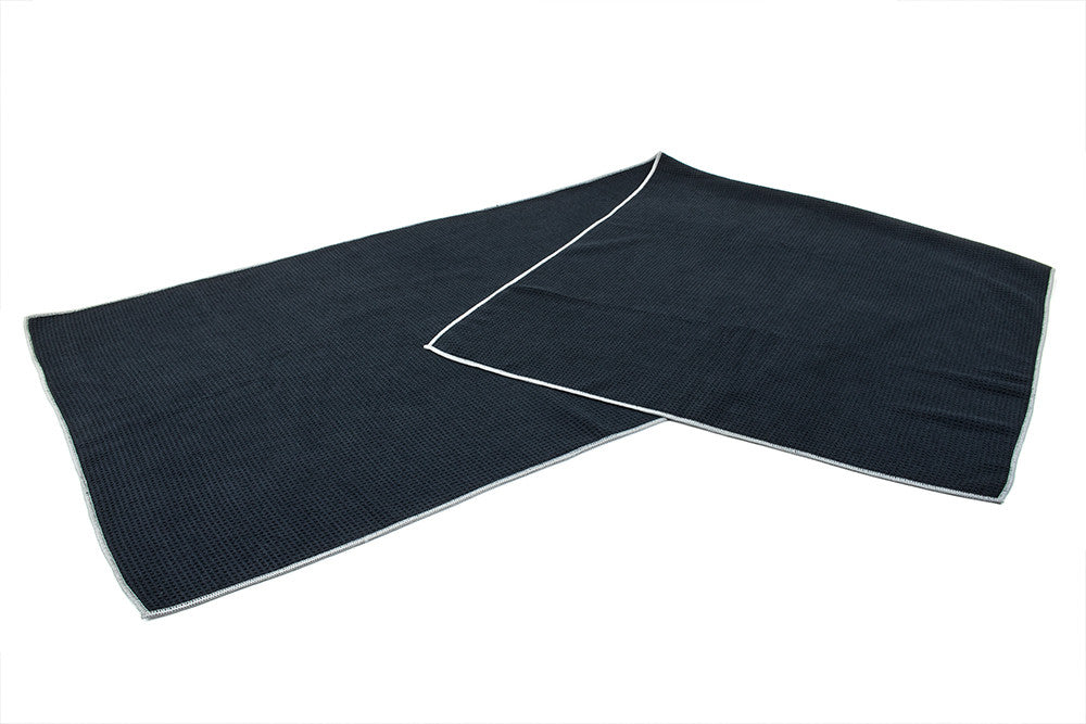 Black hot yoga mat towel, made from a high-quality, waffle weave microfiber material. Featuring a white stitching around the perimeter of the towel.